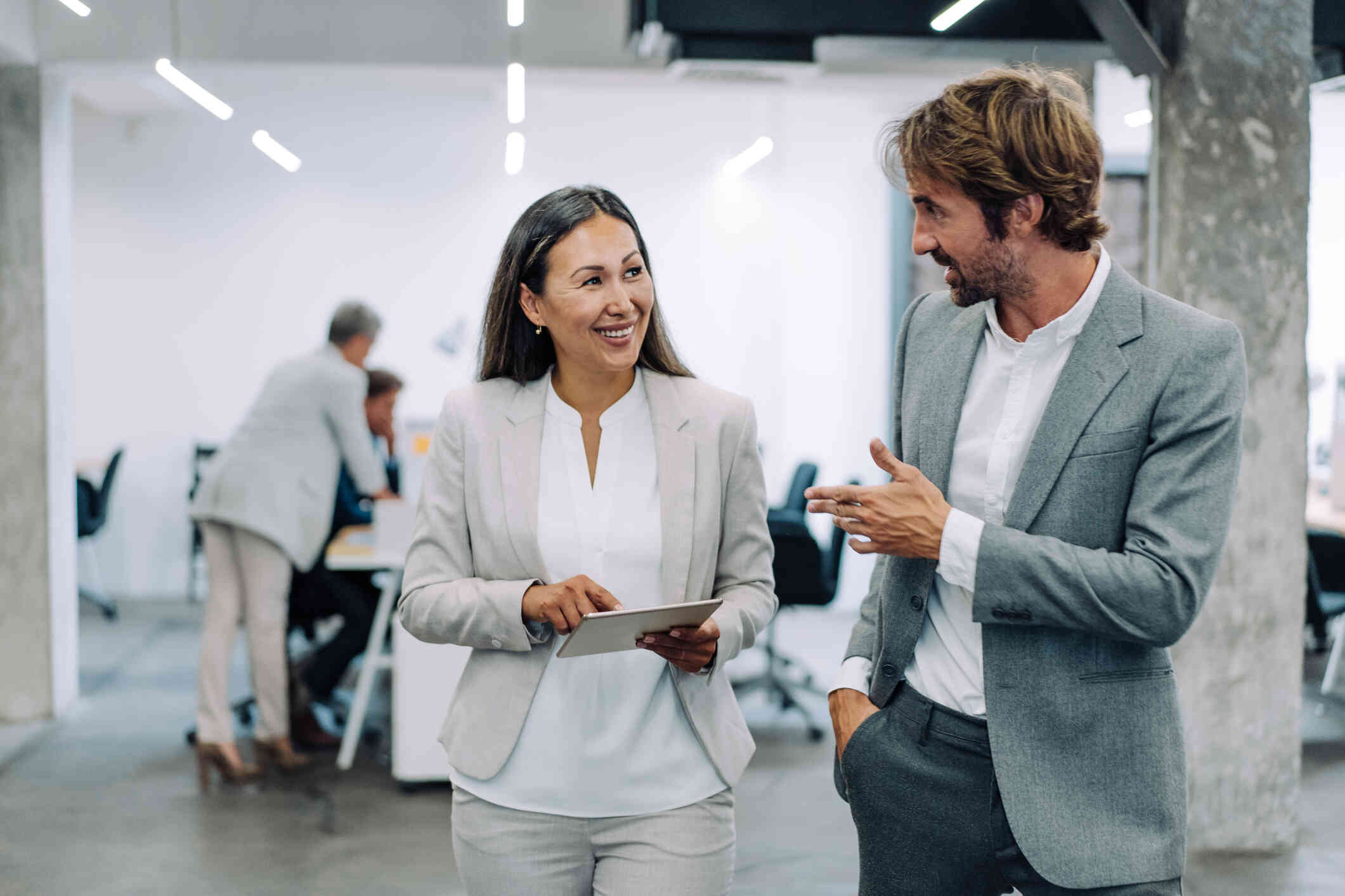 A woman in a suit smiles as she holds a tablet and walks away from a conference room next to a man in a suit who is speaking to her.